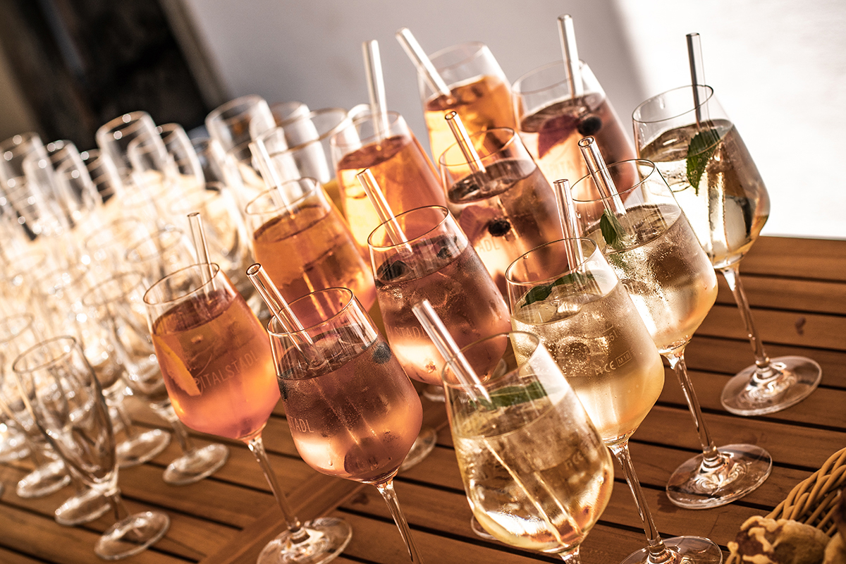 Verschiedene Aperitife auf einem Tisch, mehrere Weingläser mit Glasstrohhalmen und Eiswürfeln bestückt. Ein sommerlicher flair im Innenhof beim Sektempfang.
