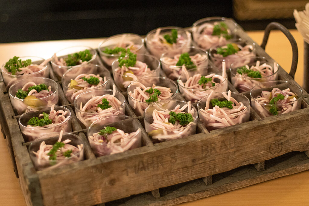 Wurstsalat am Mitternachtsbuffet in kleinen Gläsern mit Petersilie garniert. Die Gläser sind in einer rustikalen Holzkiste angerichtet.