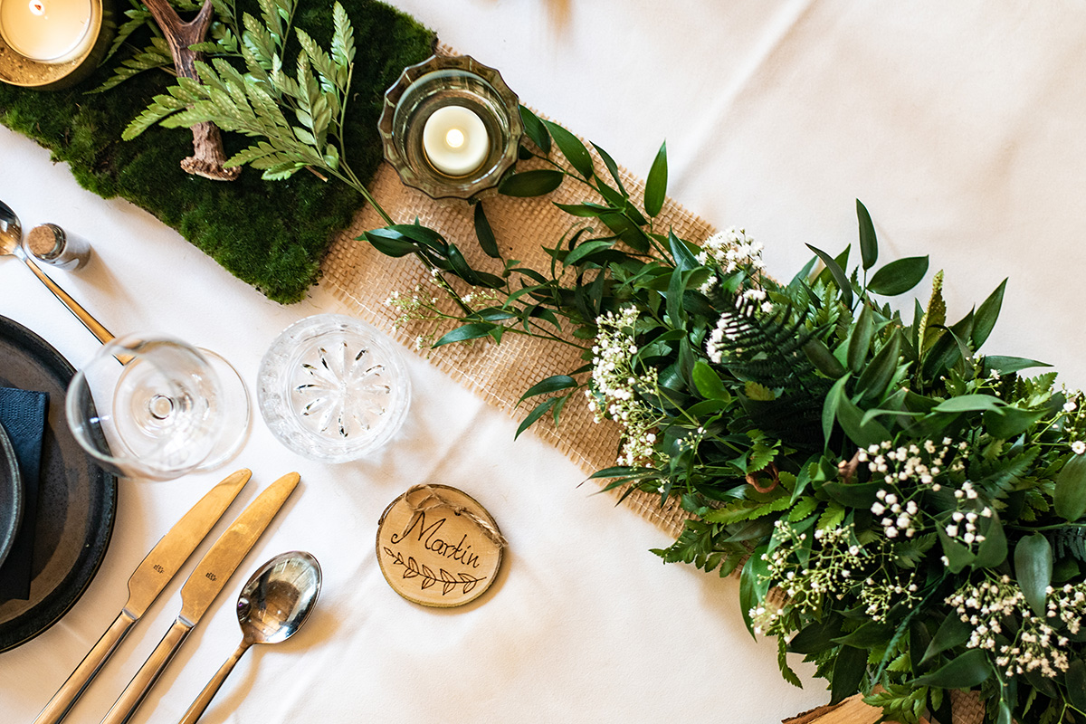 Festliche Hochzeitsdeko mit weißer Tischwäsche und Juteläufer, verziert mit grünem Blumen-Gesteck und eine kleine Holzscheibe mit Gast-Namen als Gastgeschenk. Dekoriert mit einem Rehgeweih und Moos.