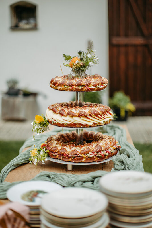 Eine dreistöckige Brezentorte verschieden belegt mit Tomate-Mozzarella, Wurst und Käse. Die Torte ist mit Blumen verziert, Sie steht auf einem Tisch im Innenhof des Spitalstadl. Auf dem Tisch befinden sich Teller und ein Boho-Stoff-Läufer als Dekoration.