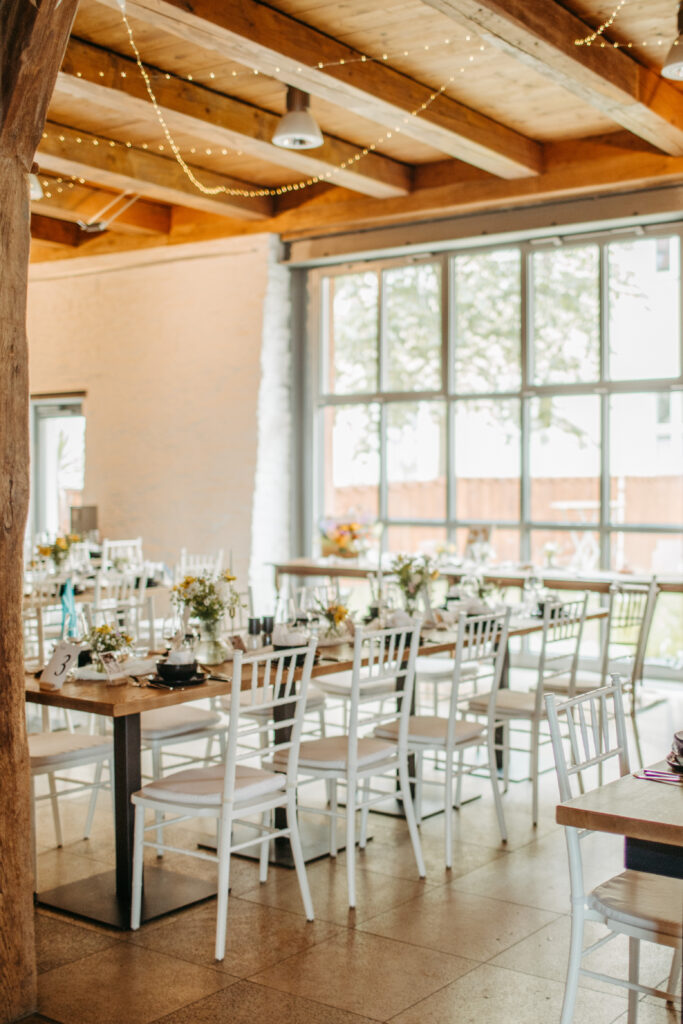 Hochzeitssaal im Spitalstadl mit dekorierten Tischen, weißen Stühlen und Lichterkette an der Decke. Das Gedeck für das Abendessen ist bereits eingedeckt. Im Hintergrund sieht man das große bodentiefe Fenster zum Innenhof