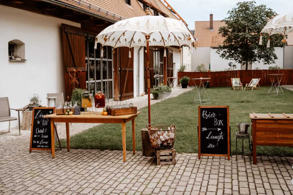 Innenhof bei einer Hochzeit im Spitalstadl mit großer Rasenfläche auf denen runde Stehtische und Liegestühle stehen, im Vordergrund ein Tisch mit einer Limobar mit zwei Sorten und Milchfläschchen als Gläser. Neben dem Tisch steht ein Balischirm, mit weißem Stoff bespannt und Goldverzierung. Daneben steht eine Holzbox als Kühlbox in der sich Bier befindet.