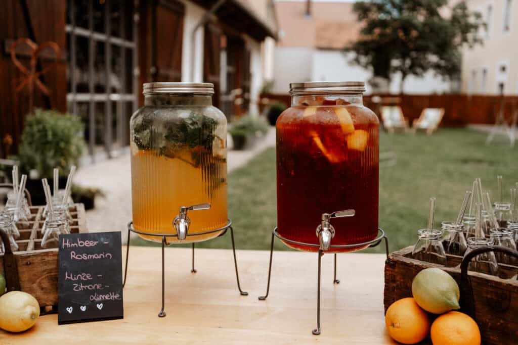Limobar mit zwei verschiedenen Sorten auf einem Tisch im Innenhof der Hochzeitslocation. Milchflaschen als Gläser mit nachhaltigem Glasstrohhalm. Dekoriert mit Orangen, Limetten und Zitronen. Die Limonaden in großen Glasbehälter mit Zapfhahn.
