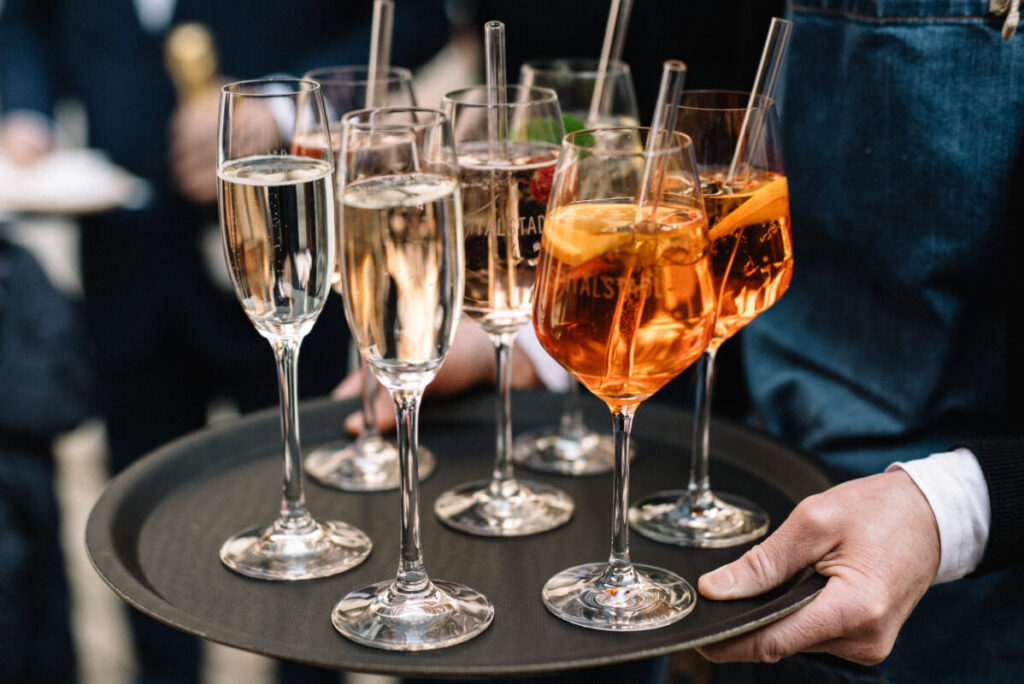 Servicekraft mit Tablett auf dem Sekt und Aperitive stehen, Aperol im Weinglas mt Orangenscheibe und nachhaltigem Glasstrohhalm.