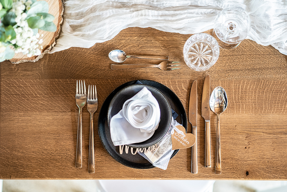 Tischgedeck für das Abendessen auf einem Massivholz-Eichentisch. Messer, Gabel, Löffel, Dessert-Löffel und Dessert-Gabel. Ein Nachtmann-Kristallglas und ein Weinglas. Schwarzes Steingutgeschirr mit Unterteller und Suppenschüssel, darin eine Stoffservierte in Rosenform.