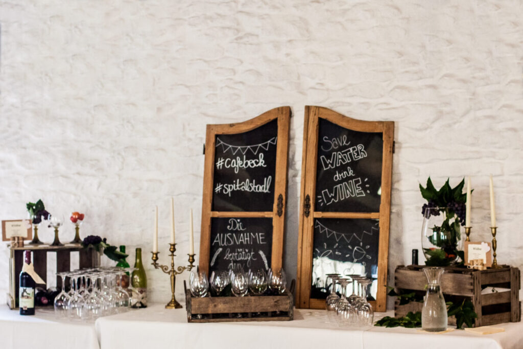 Weinbar im Hochzeitssaal, Schranktürchen mit Beschriftung zur Dekoration und Weingläser, Kerzenhalter und Weinblätter dekoriert auf einem Tisch.