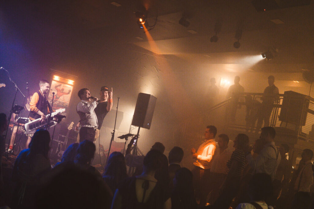 Die Band steht auf der Bühne im Kinosaal, der Bräutigam singt mit. Im Vordergrund Gäste auf der Tanzfläche die feiern.