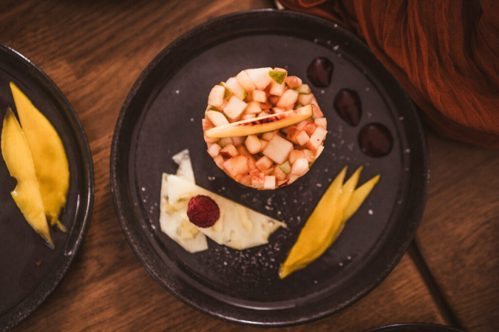 Obstsalat aif einem schwarzen Dessertteller. Mangostreifen, Ananas & Himbeere auf dem Teller angerichtet mit Saucenklecksen.