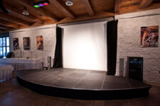 Bühnenpodest im Eventsaal mit Vorhang, Leinwand und Beamer für eine Diashow. Theaterbeleuchtung und Farbstrahler.