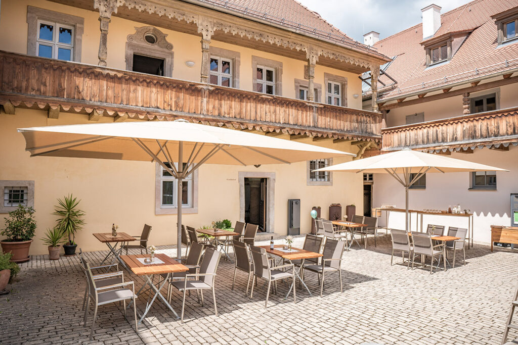 Gepflasterte Fläche im Innenhof des Spitalstadls, zwei große Sonenschirme in beige, darunter Gartenbestuhlung für ca. 40 Personen. Im Hintergrund der Holzbalkon des alten Spitalstadl-Gebäudes. Am Bildrand zwei Stehtische.